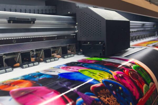 Large-format printer with black printer head shown operating and producing a large glossy banner with assorted colorful images on what appears to be vinyl.