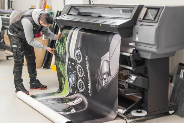 A man in a puffy vest and camouflage pants works with a printer to produce a large image of a dashboard.