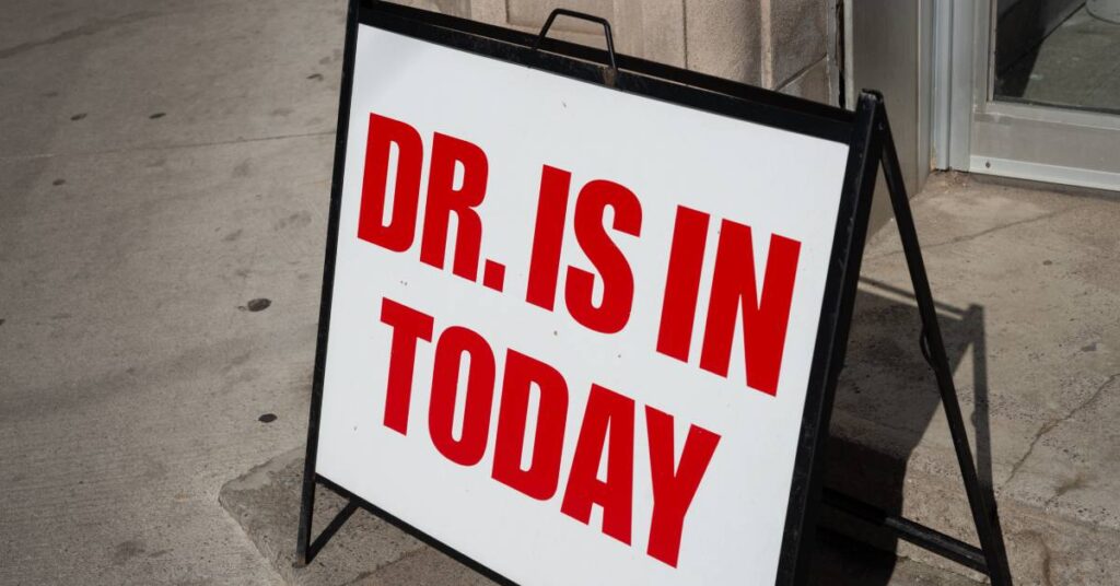 A large white a-frame sign that says "Dr. Is In Today" rests on the sidewalk in front of a door on a very bright day.