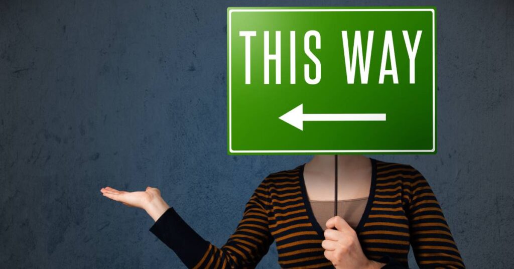 A woman holds a green directional sign in front of her face that says "THIS WAY" with an arrow pointing left.