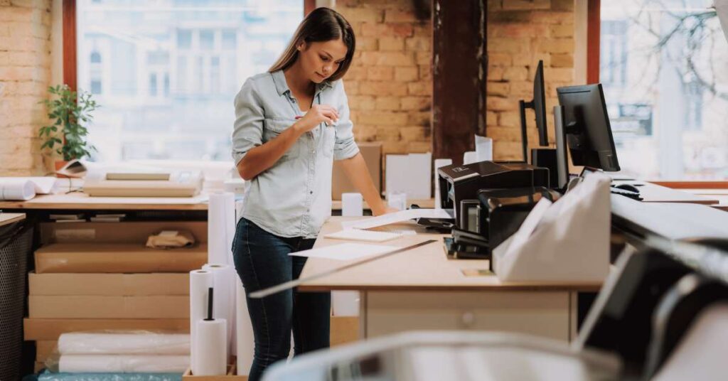 A woman is seen in an office setting observing a stack of papers just delivered by a printer on her desk.