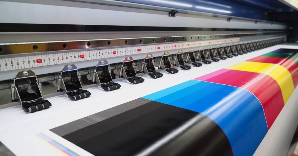 A wide-format printer, up close, prints a big sheet colored with blocks of black, red, blue, yellow, green, and more.