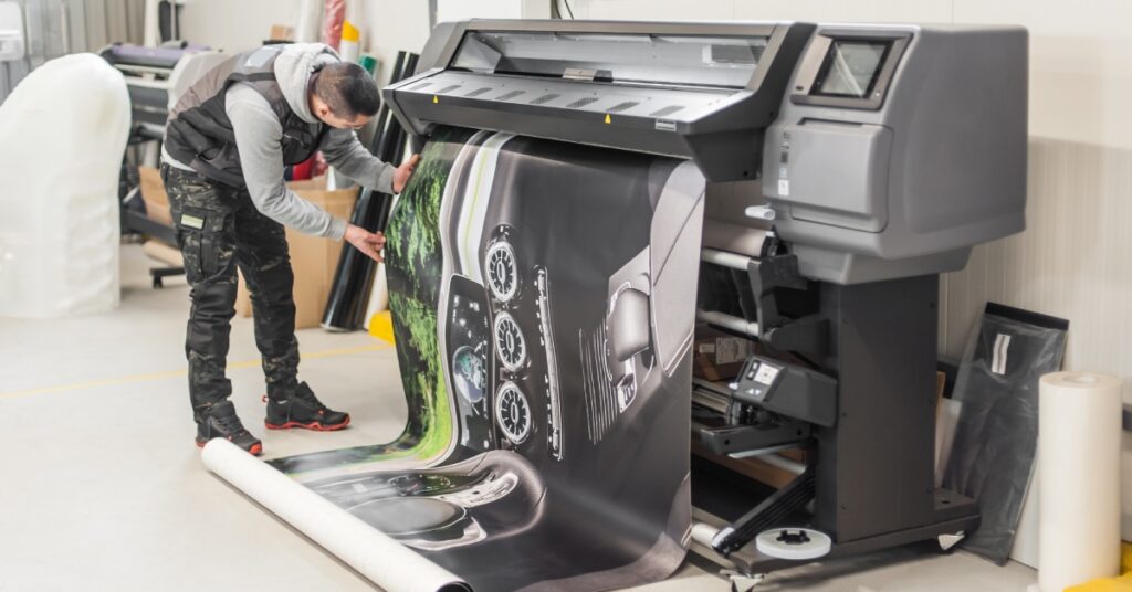 A technician in a vest and camouflage pants carefully inspects a printed sheet emerging from a plotter printer.