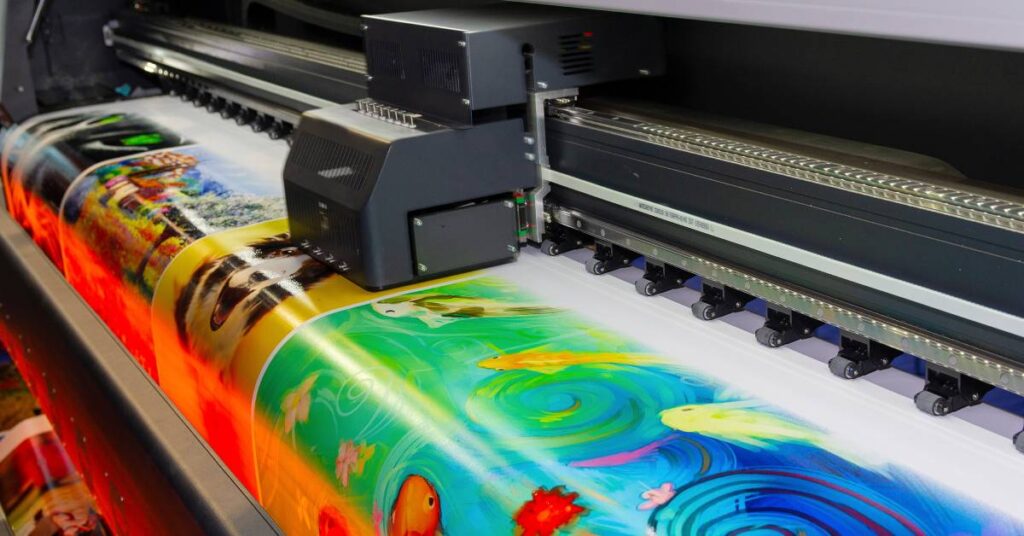 A large-format printer produces several large photographs in assorted bright colors on a wide sheet of media.