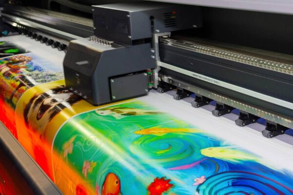 A large-format printer produces several large photographs in assorted bright colors on a wide sheet of media.