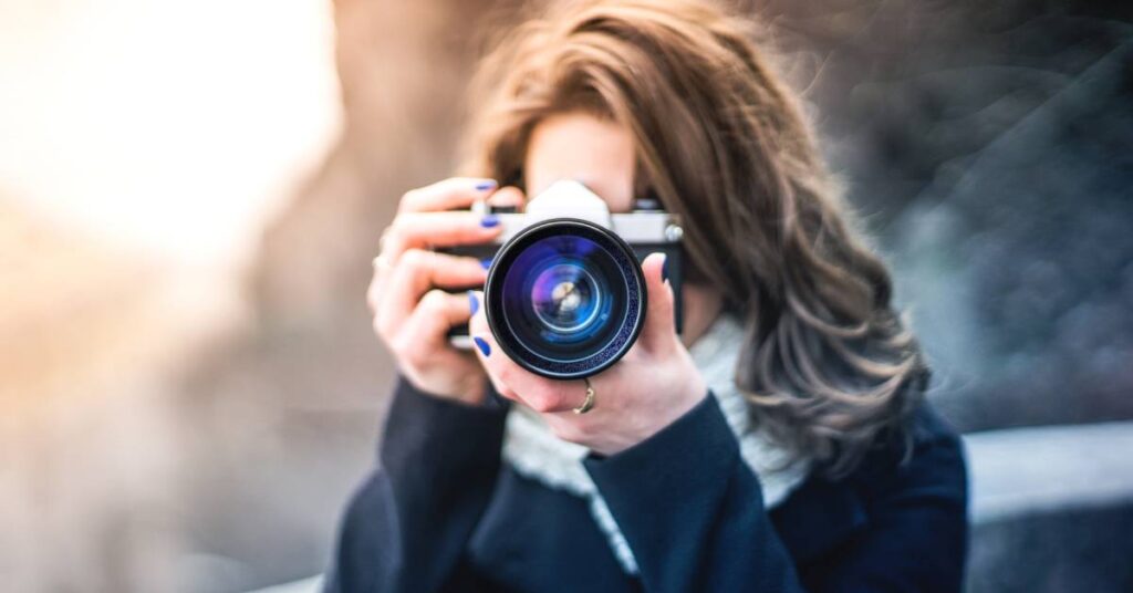 A young woman wearing a scarf and black shirt points a camera at the viewer, as if to take their picture.