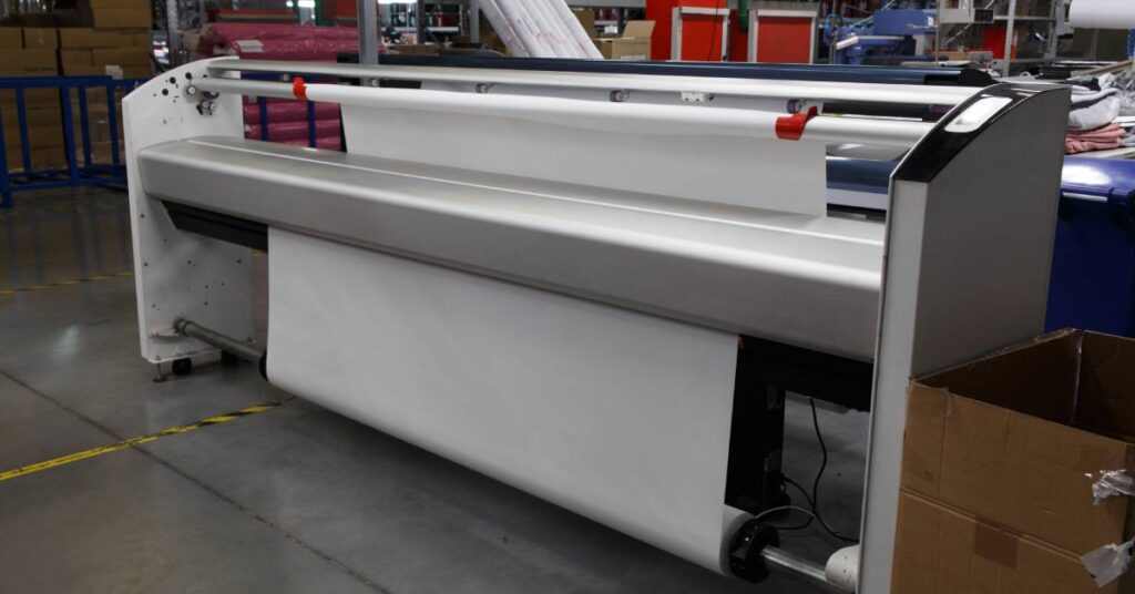 A wide-format plotter printer sits in a print shop or factory setting equipped with a large roll of print media.