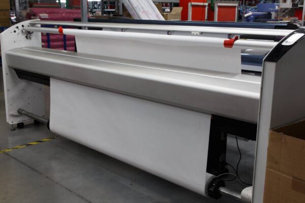 A wide-format plotter printer sits in a print shop or factory setting equipped with a large roll of print media.