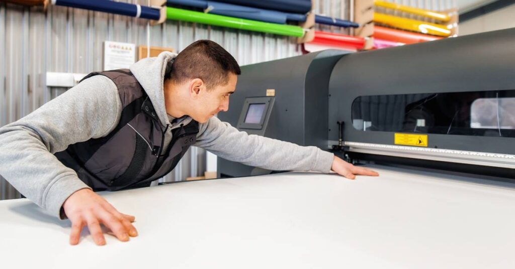 A man in a vest and hoodie is aligning a large sheet of paper in a industrial plotter printer device.