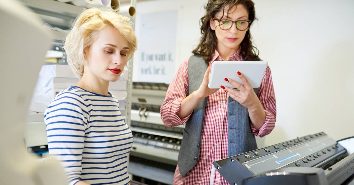 Two women in a printing workshop make adjustments to the settings on their plotter machine for their project needs.