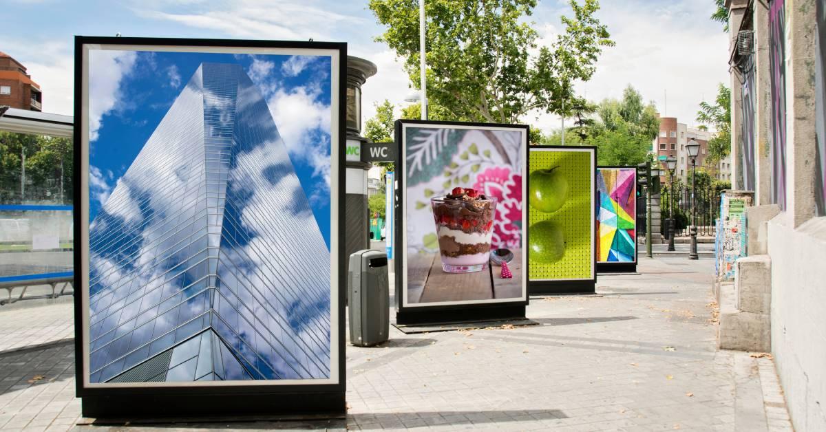 An empty city street with four large billboards with different images like ice cream, a skyscraper, apples, and a mosaic.