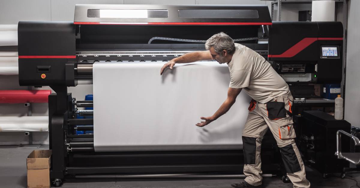 A technician loads a large blank paper into a plotter printer. There are several more rolls hung on the wall behind them.