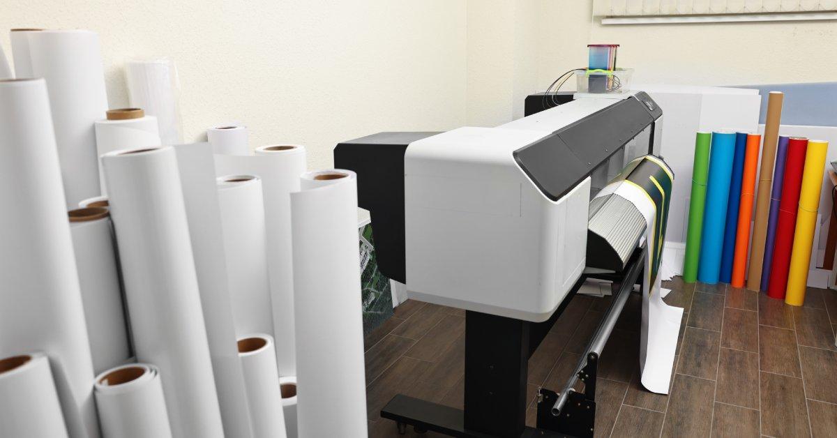 A room with a wide format printer and several rolls of white and colored paper. A large sheet hangs from the tray.