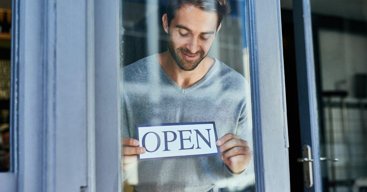 A shop owner places an "open" sign on the window of a business front. The door next to him is cracked open.