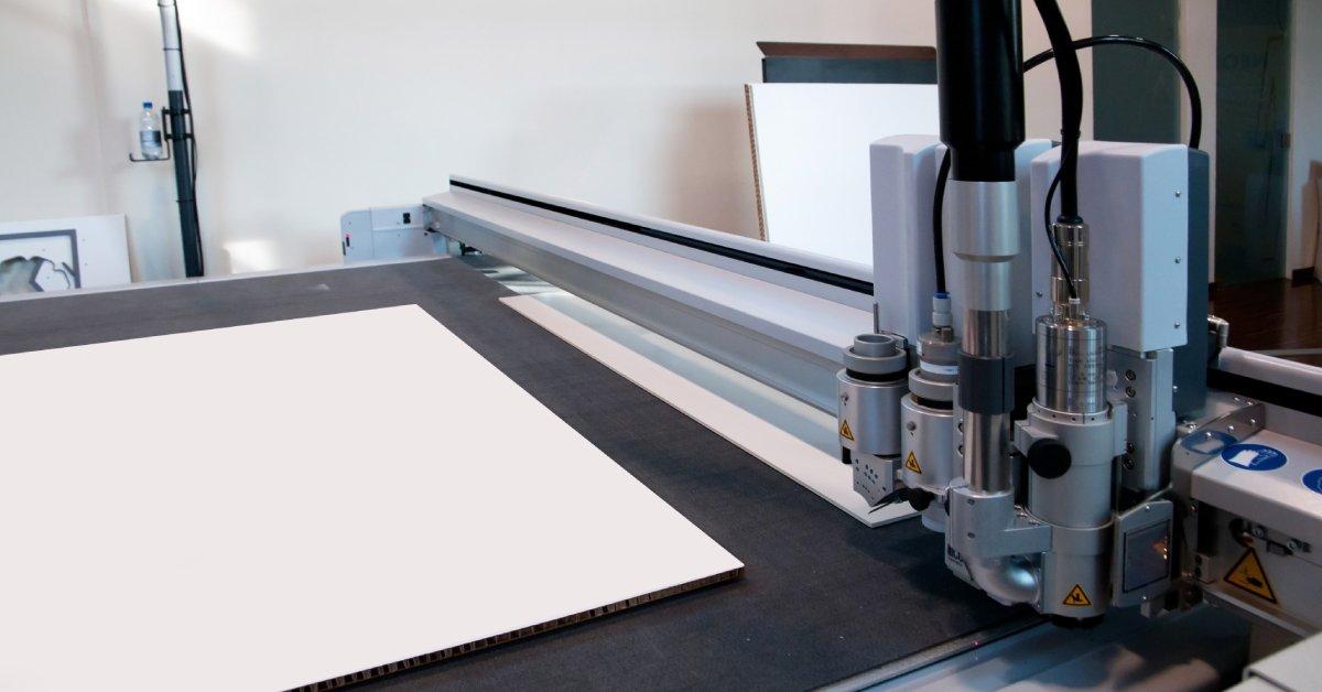 A plotter with a blank corrugated print on its flat surface. A black cable connects the printing mechanism to the machine.