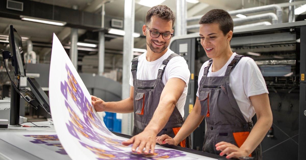 Two graphic designers smile as they inspect the quality of a printed deliverable in a modern print shop.