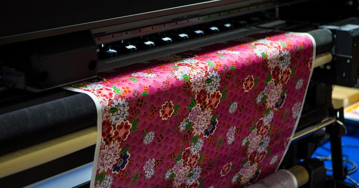 A wide-format printer uses a roll of textile material to produce a colorful red and pink floral design.